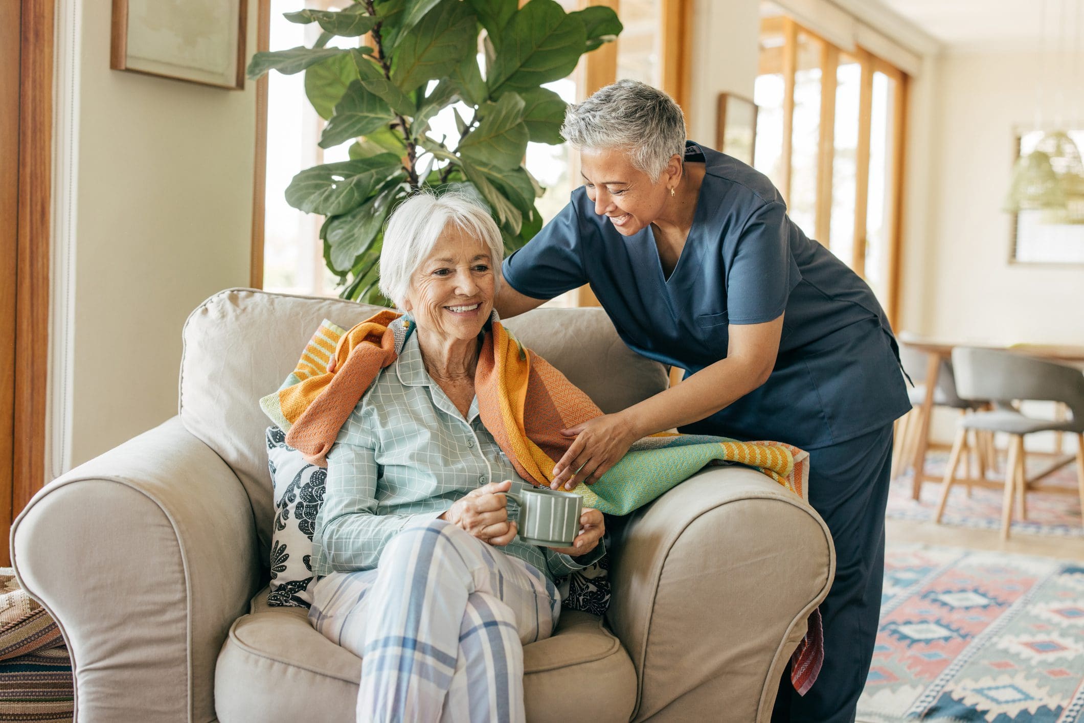 Elderly woman smiling while nurse adjusts her scarf at home.