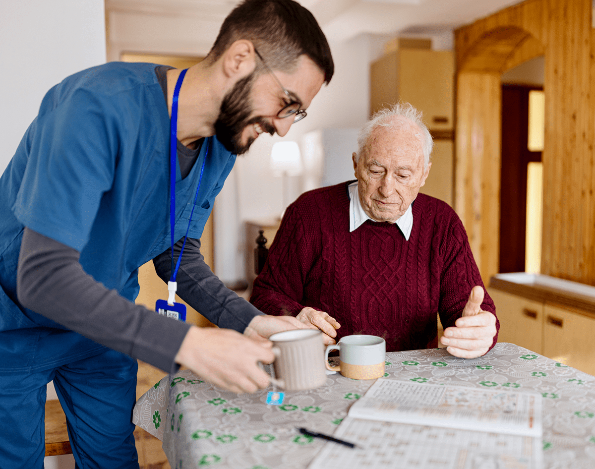 A caregiver helps an elderly man with his meal at a table.