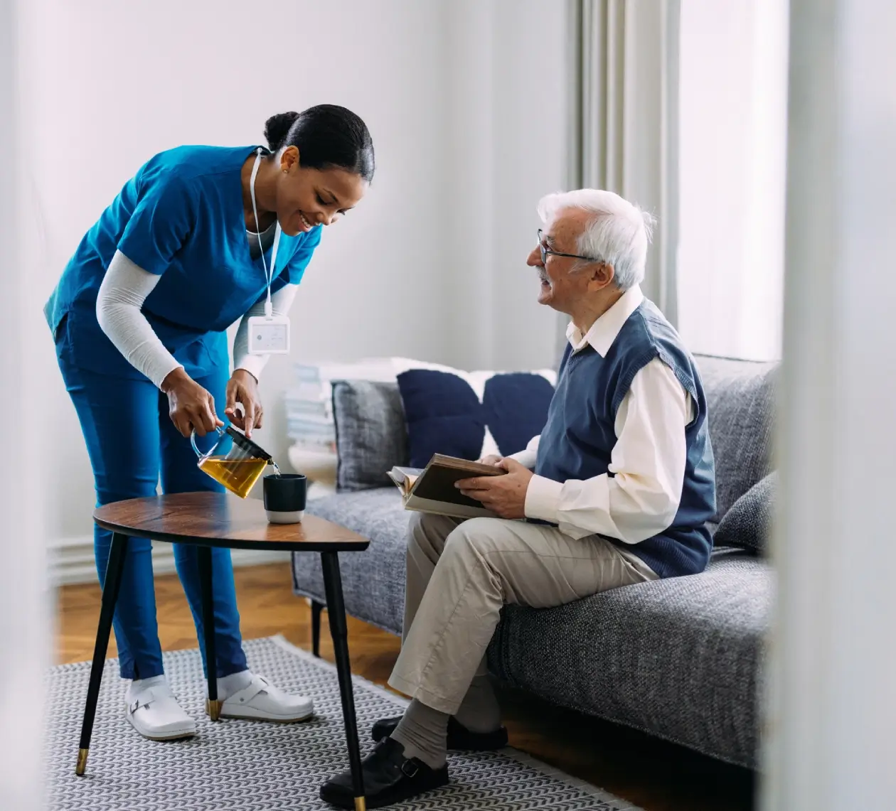 A caregiver serving tea to an elderly man in a cozy living room.